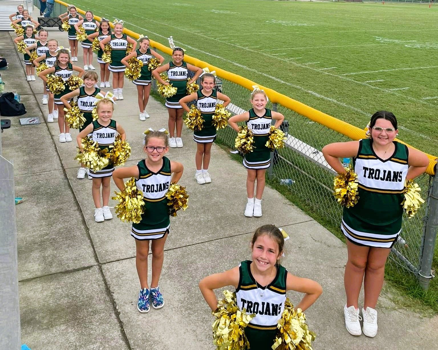 Cheerleaders in green and gold uniforms holding gold pom-poms line up on a concrete surface next to a fence.