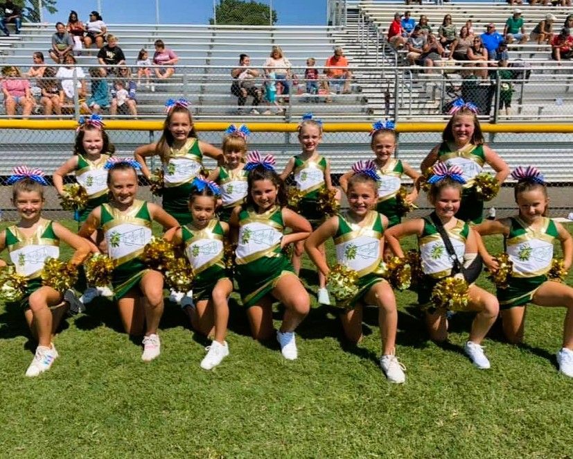 Youth cheerleaders in green and gold uniforms on a grassy field, posing with pom-poms.
