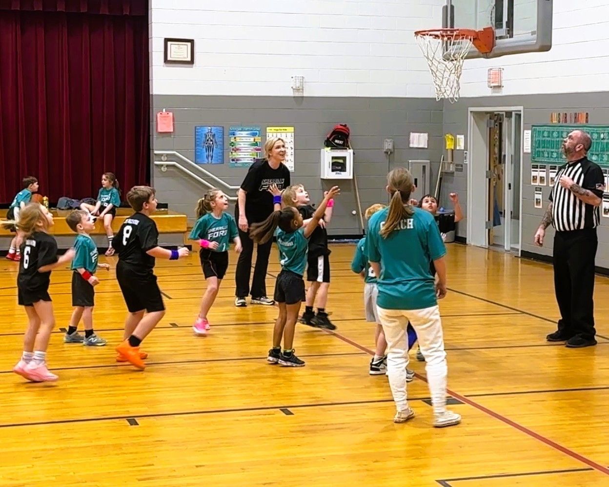 Youth basketball game in a gymnasium; children in teal and black uniforms play, referee and coaches observe.