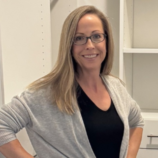 Woman in glasses and cardigan smiling in front of white shelving.