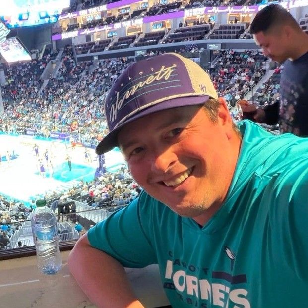 Man smiling, wearing a Hornets hat and shirt, at a basketball game.
