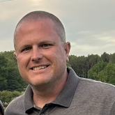 Man with short hair smiles outdoors, wearing a gray polo shirt, trees in the background.
