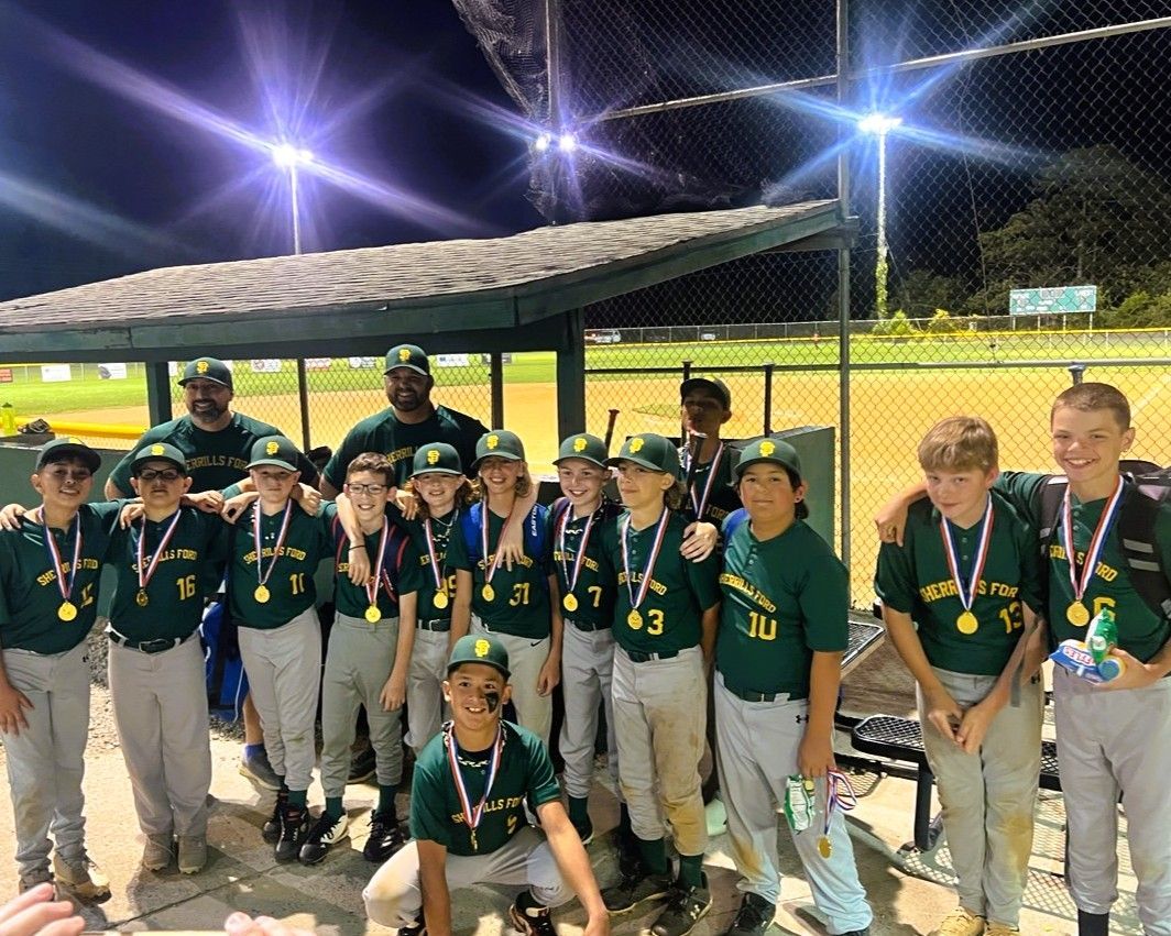 Youth baseball team in green uniforms and medals, posing under dugout roof after a game.