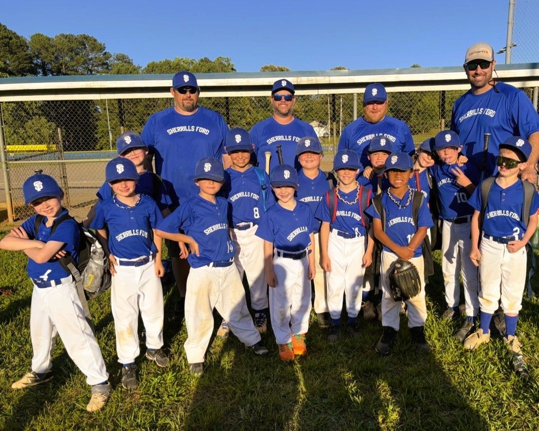 Youth baseball team in blue uniforms stands on a field. Coaches in back, players with hats and bats.