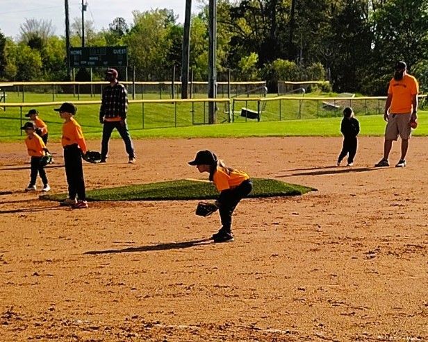 Children in orange shirts and black pants practice baseball fielding on a dirt field.
