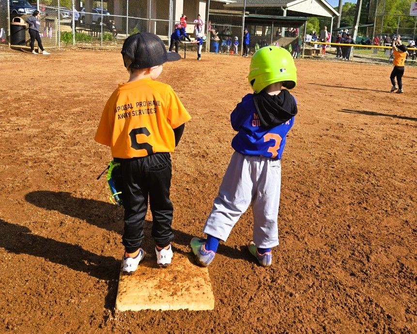Two young baseball players on a dirt field, one wearing orange and the other blue, standing on a base.