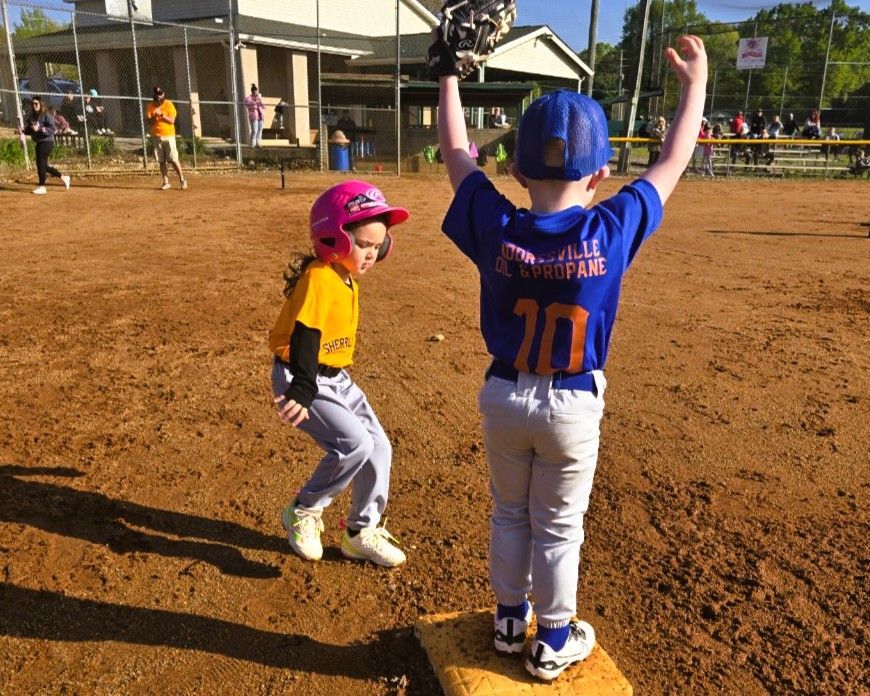 A child in blue baseball uniform with arms raised celebrates, while another in pink helmet looks on. Baseball field.