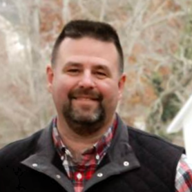 Man smiling outdoors, wearing a black vest and plaid shirt, with trees in the background.