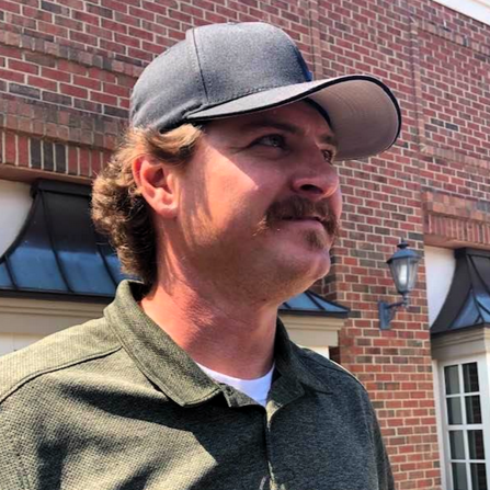 Man with a mustache wearing a gray cap and green shirt, standing outdoors in front of a brick building.