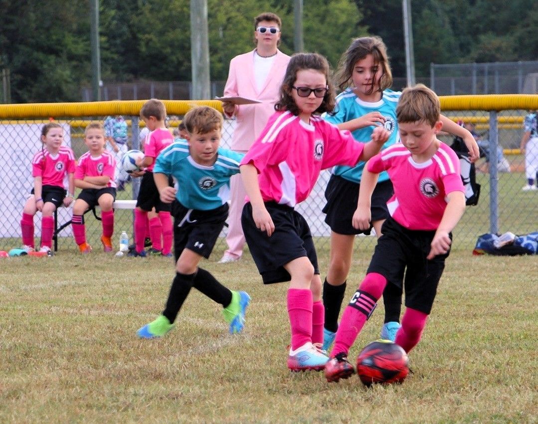 Children playing soccer, one kicking ball, others nearby, coach in pink blazer watches from the background.