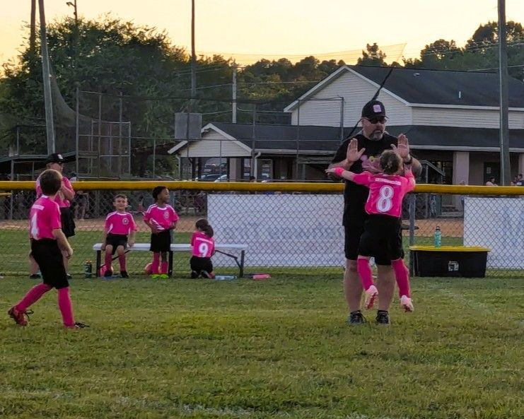 Youth soccer team in pink uniforms celebrating with their coach on a grassy field.