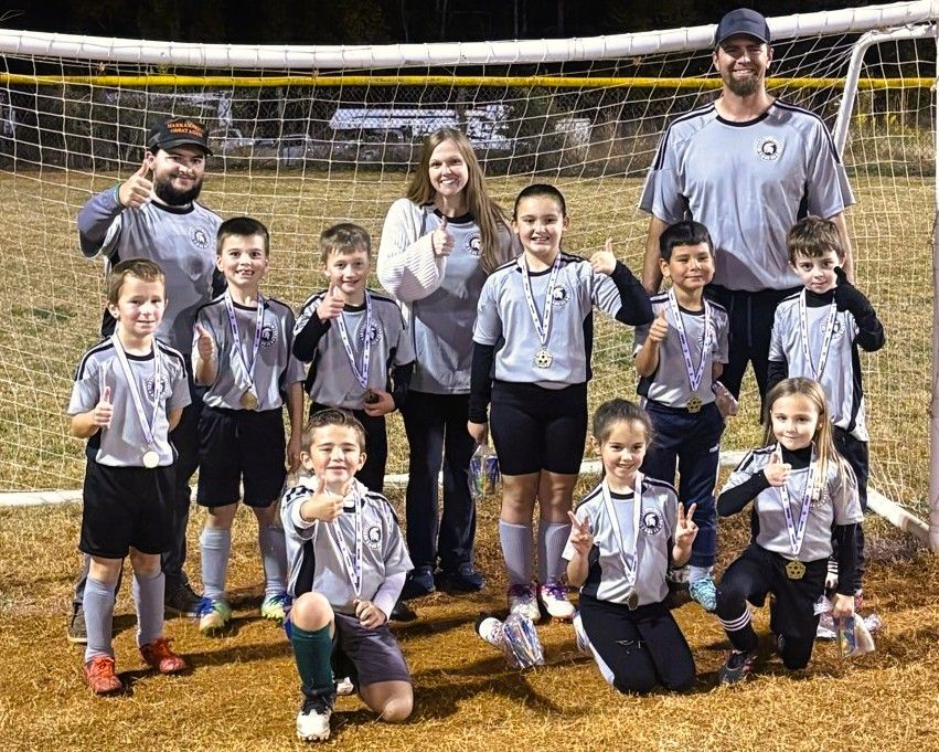 Soccer team posing with coaches on a field, all wearing gray jerseys and giving thumbs-up.