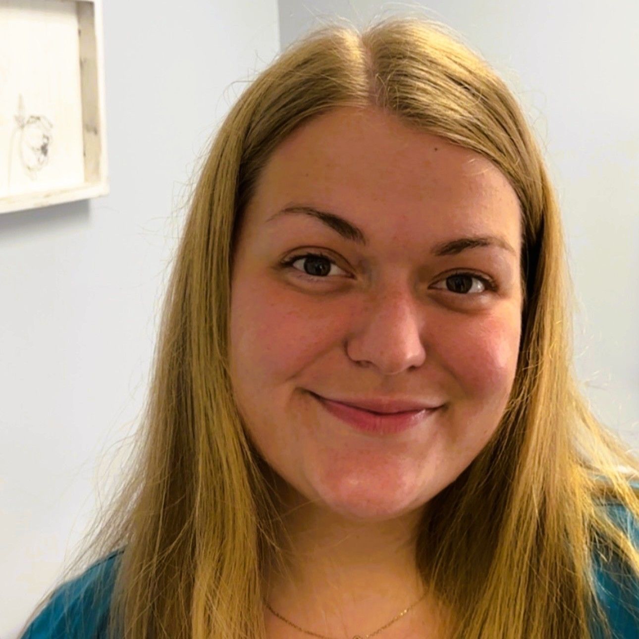 Woman with long blonde hair smiles, wearing a teal top, indoors against a light blue wall.