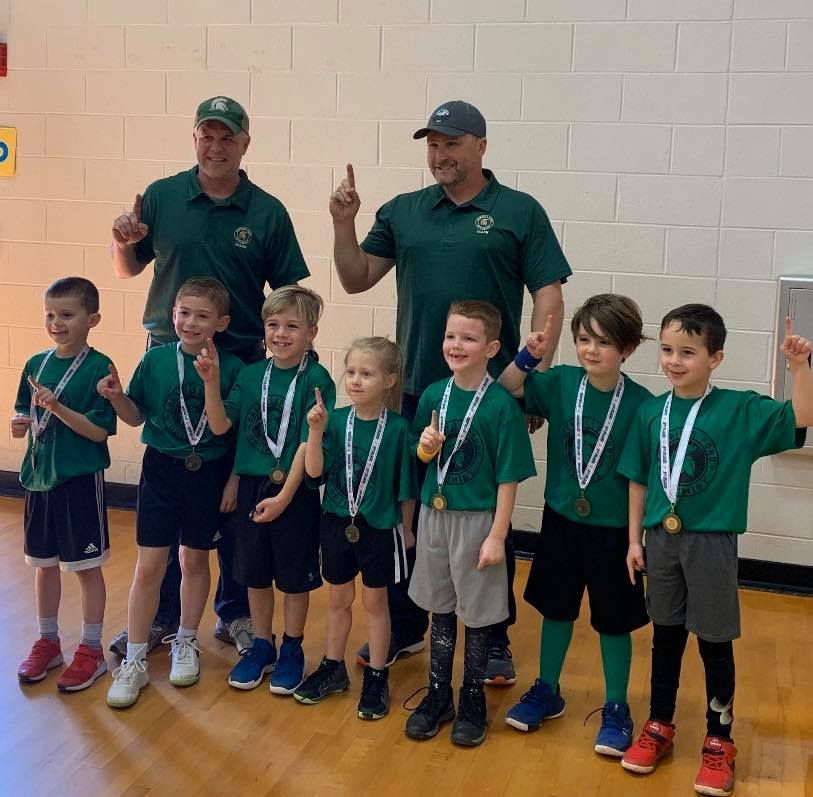 Youth basketball team and coaches wearing green shirts and medals, raising index fingers in a gymnasium.