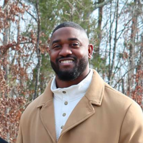 Man smiling outdoors, wearing a tan coat and white sweater.