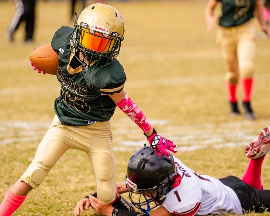 Youth football player running with the ball, dodging a tackled opponent on a grassy field.