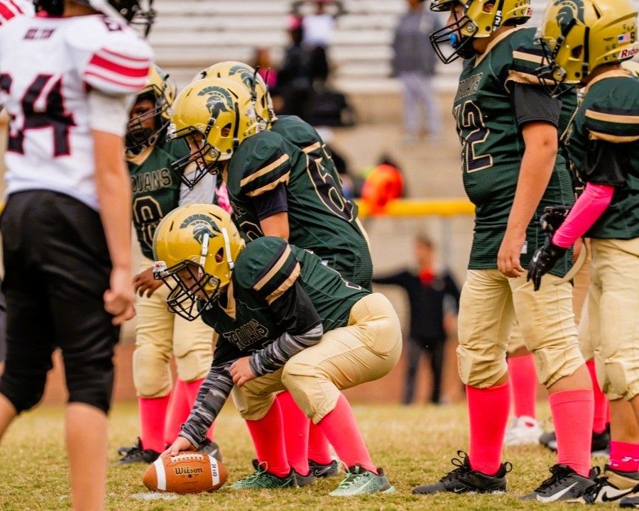 Youth football players huddle, preparing for a play on a green field. Players wear green and gold uniforms, pink socks.