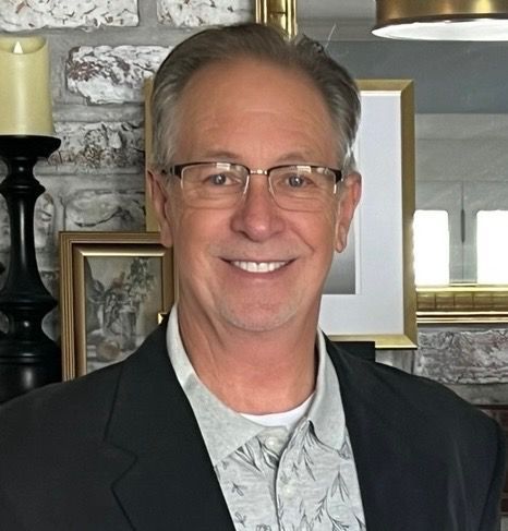 Man with glasses smiles, wearing a blazer over a patterned shirt, in front of a brick wall, art, and a candle.