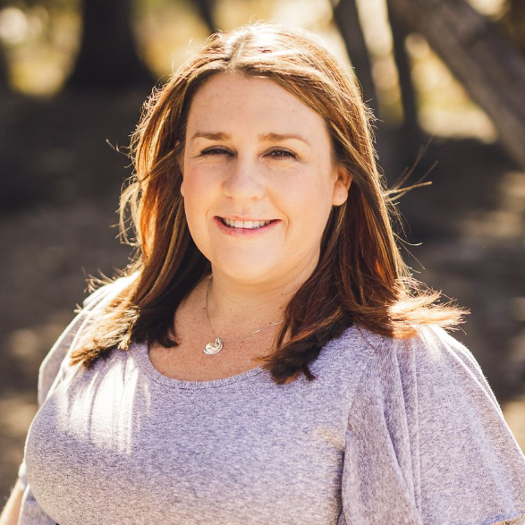 Woman with brown hair smiles, wearing a grey shirt, outdoors.