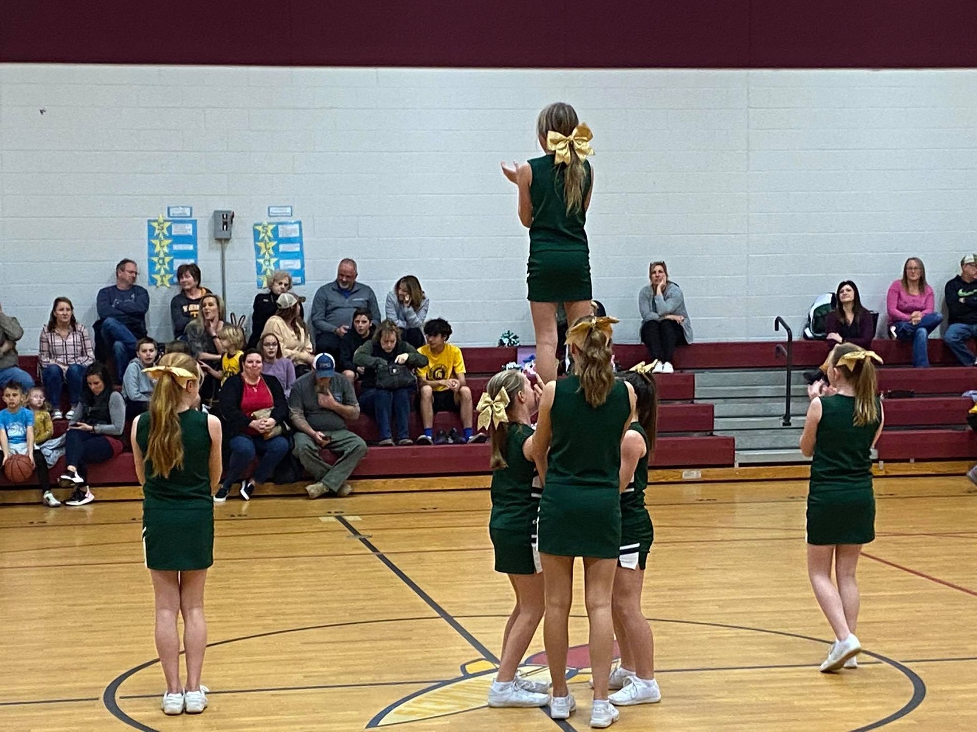 Cheerleaders in green uniforms perform a stunt on a basketball court, watched by an audience.