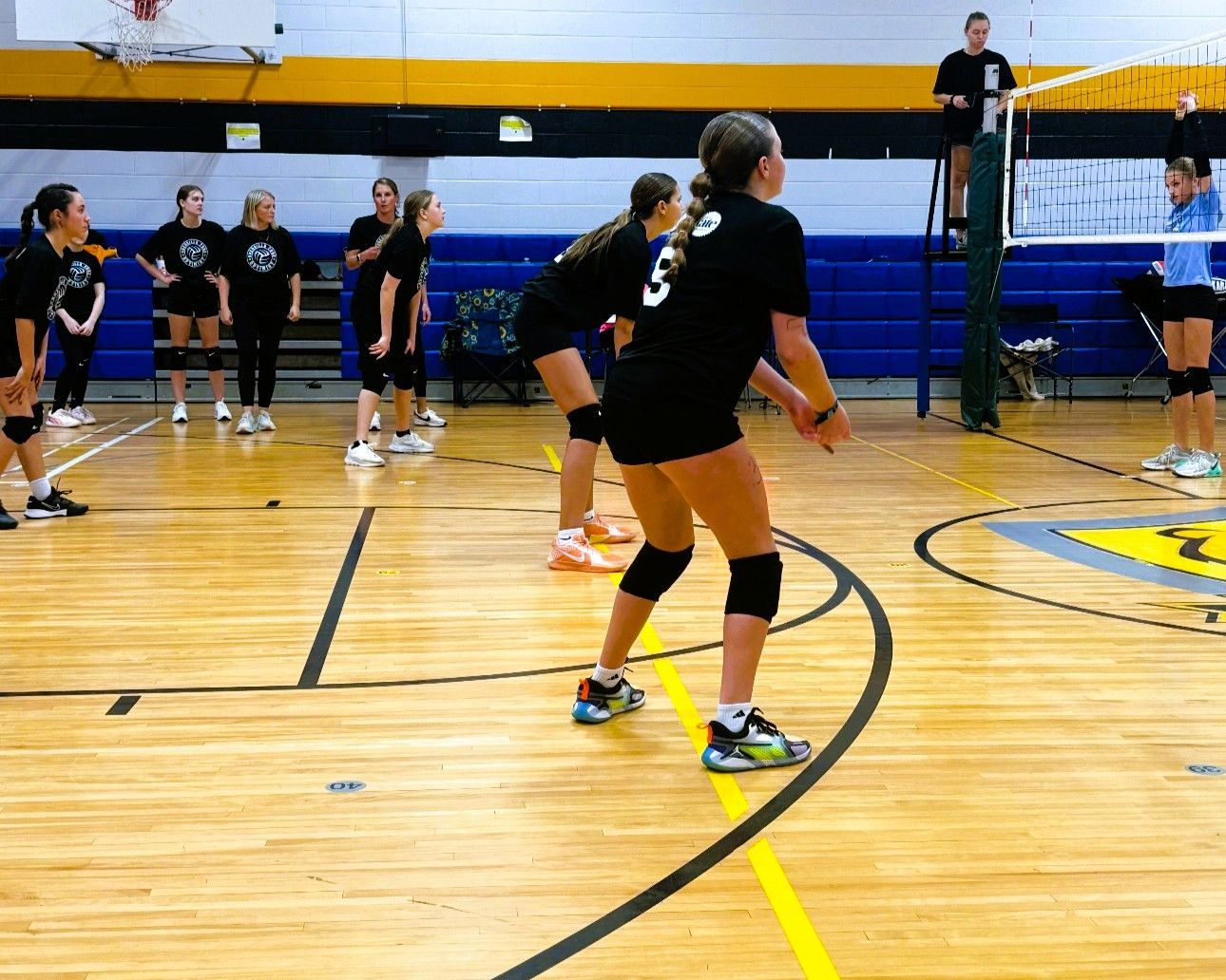 Volleyball players in black uniforms, practicing in a gym. Player in focus is in receiving stance.