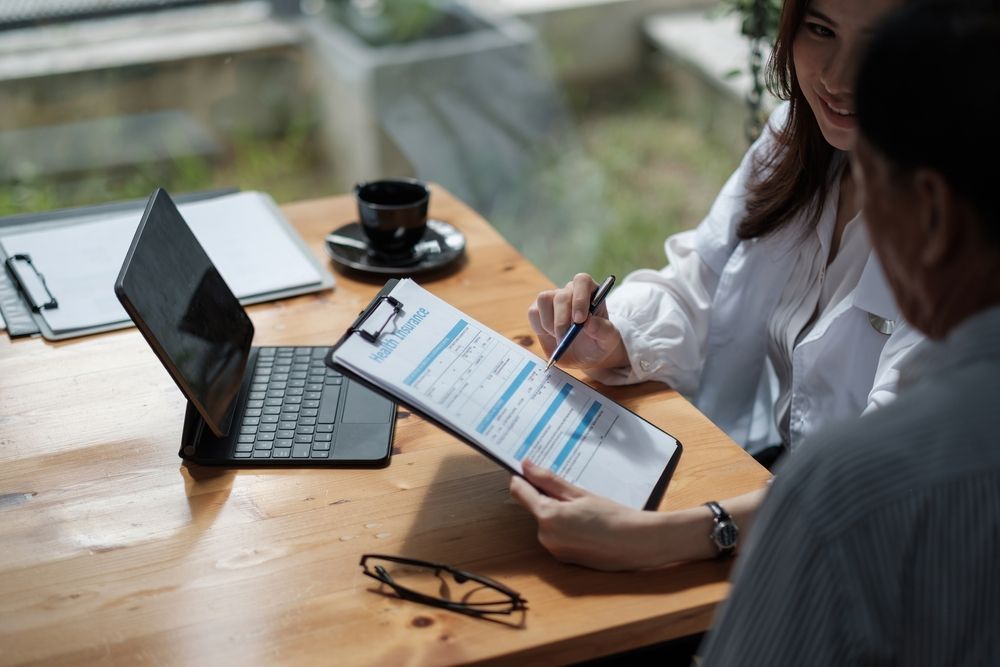 Woman pointing to paperwork for a man at a table with a laptop, coffee cup, and glasses.