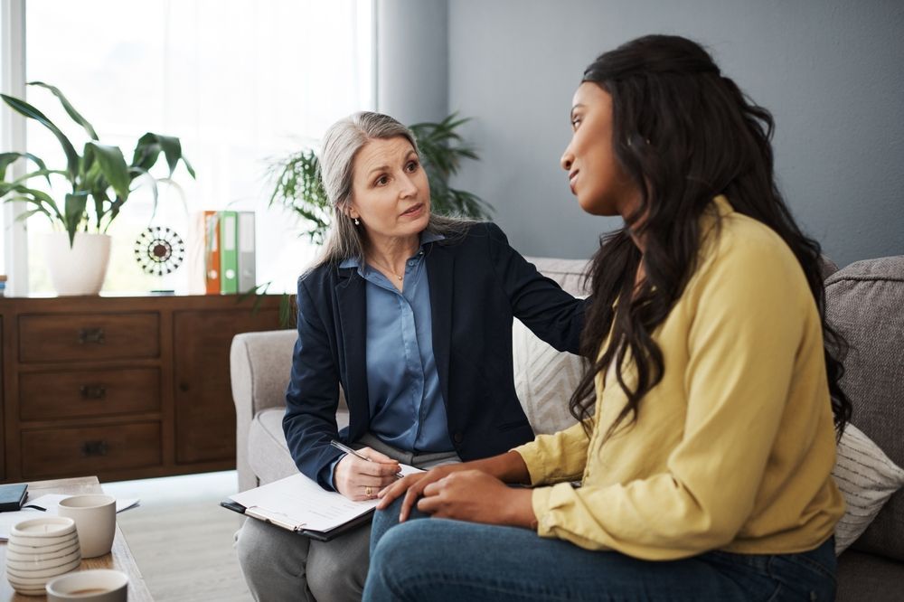 A therapist comforts a Black woman, writing notes. Both are seated on a couch in an office.