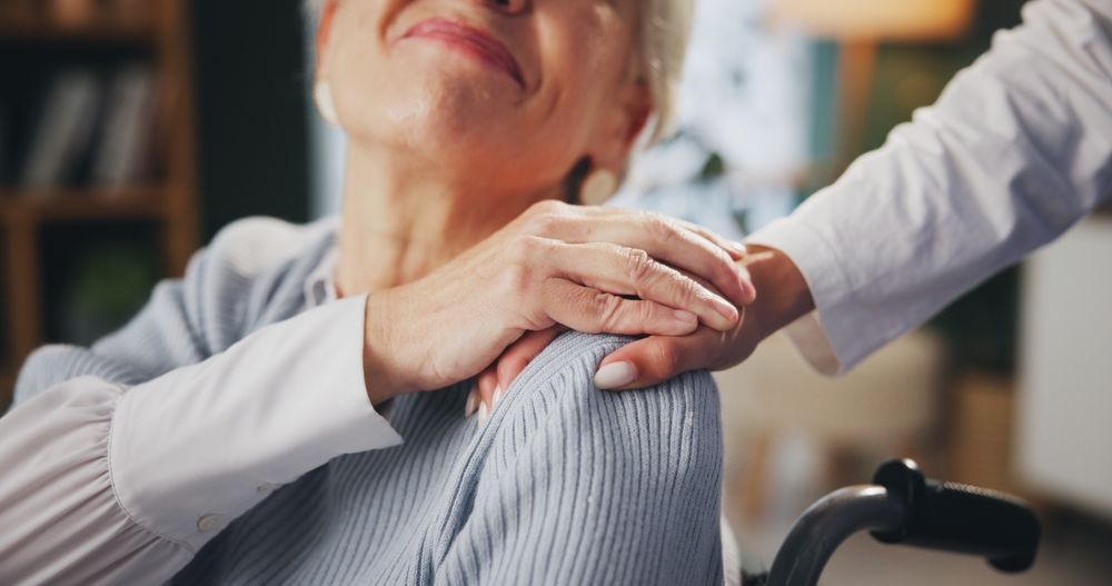 Older woman in a wheelchair, smiling, hand on shoulder by someone in a white coat indoors.