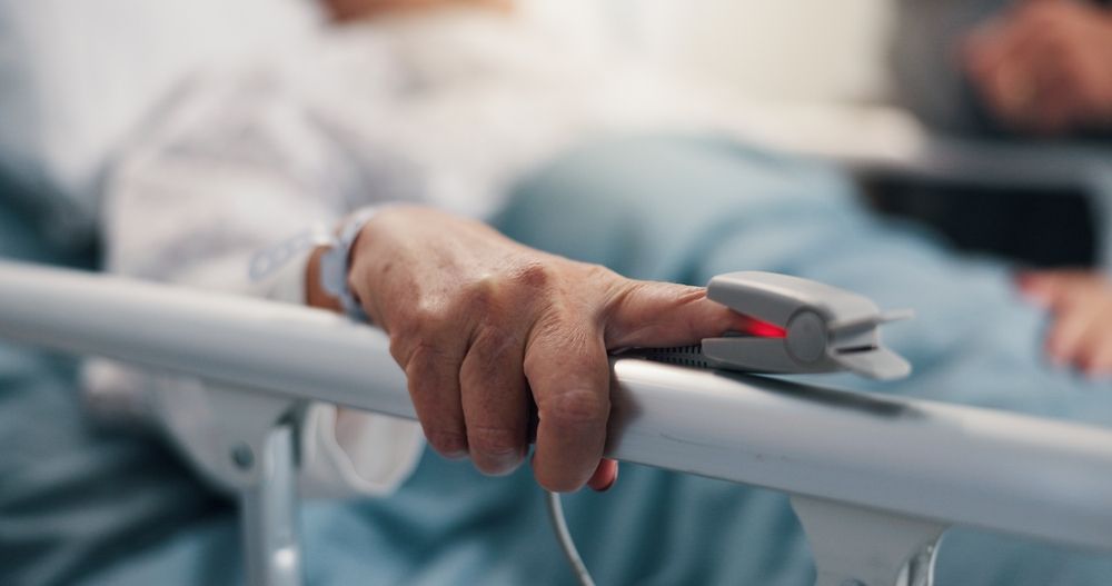 Person in hospital bed, hand on rail with pulse oximeter clipped to finger.