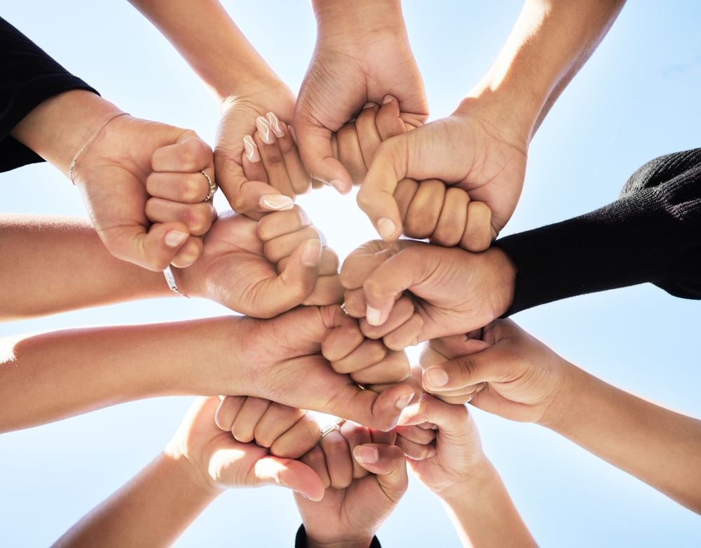 Hands joined together in a circle, fists touching, viewed from below against a blue sky, symbolizing unity.