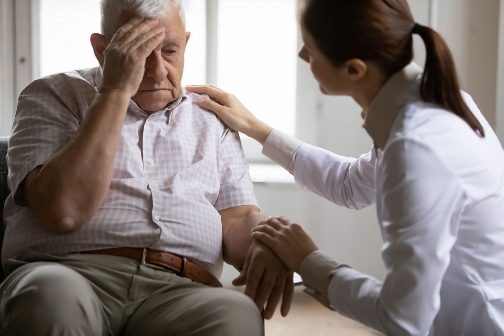 An elderly man with hand on forehead, looking distressed, is comforted by a woman with hand on his shoulder.