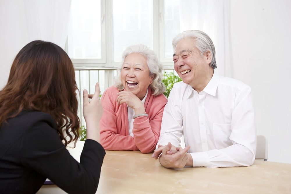 A businesswoman gestures to laughing elderly couple at a table in a bright room.