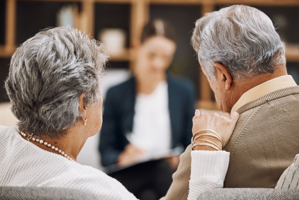 Elderly couple in therapy session, facing away, listening to blurred therapist.