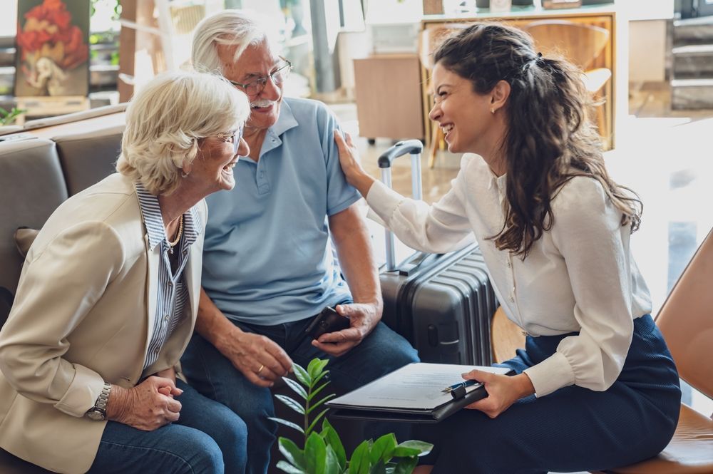 Woman assisting senior couple; smiles, paperwork, luggage in lobby.