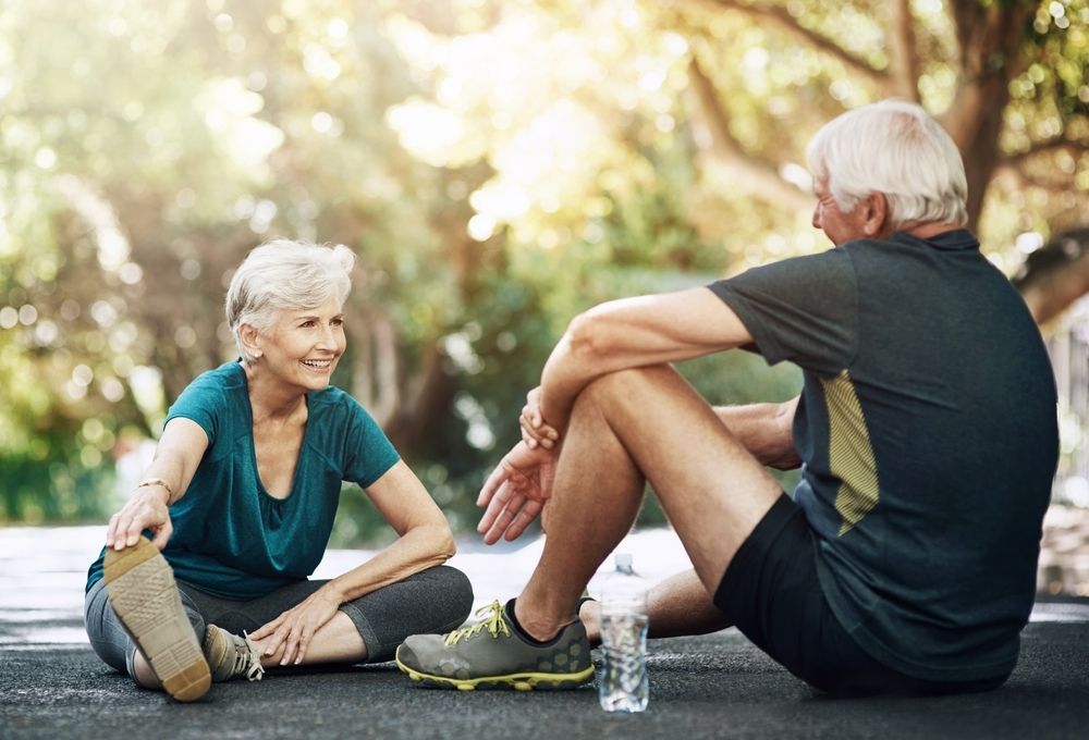 Elderly couple resting on a path after exercising outdoors. Woman stretches; man sits with water bottle.