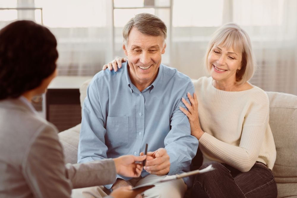 Senior couple smiling, sitting with a consultant. Man holds pen, woman's hand on his shoulder. Indoors, natural light.