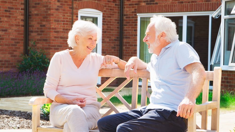 Senior couple smiling and talking while seated on a wooden bench outside a brick building.