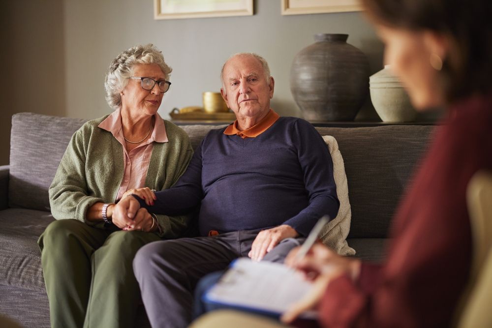 Elderly couple holding hands on a couch, facing a person taking notes; inside a home.