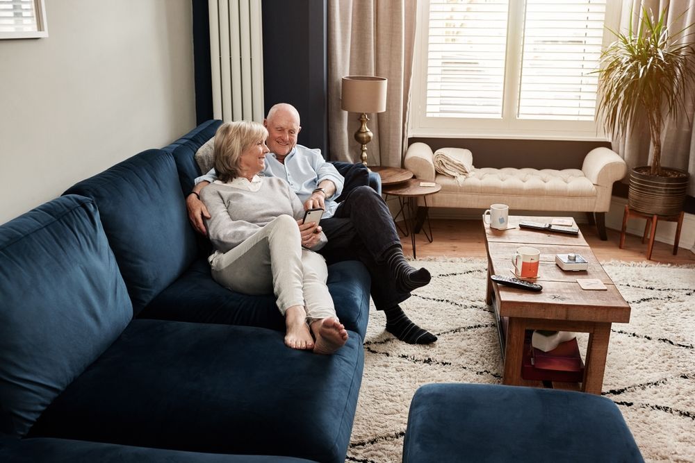 Elderly couple cuddled on a navy sofa, smiling in a well-lit living room with neutral colors.