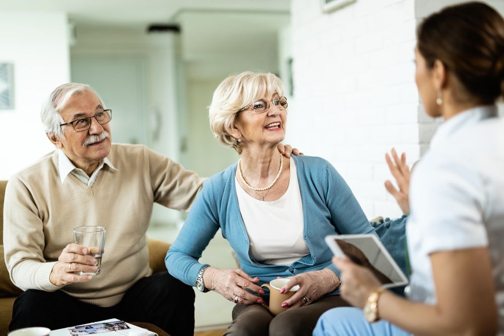 Senior couple conversing with a healthcare worker in a living room; the woman holds pills.