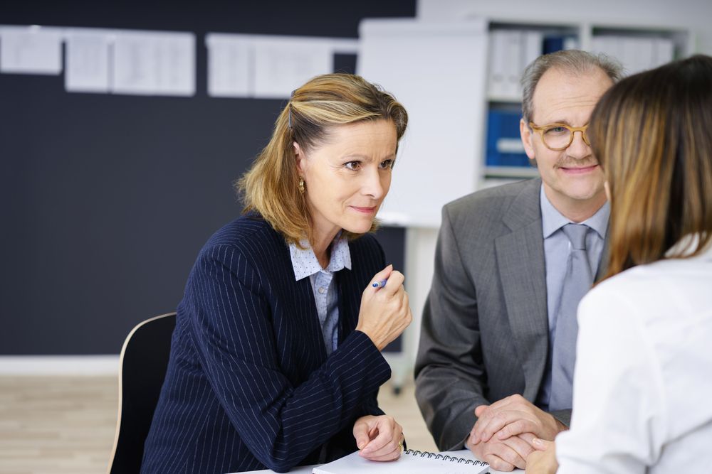 Three businesspeople in a meeting, one woman speaking, two looking at her, office setting.
