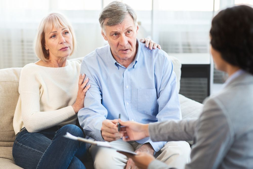 Senior couple in therapy, discussing with counselor. Sad expressions, holding a pen.