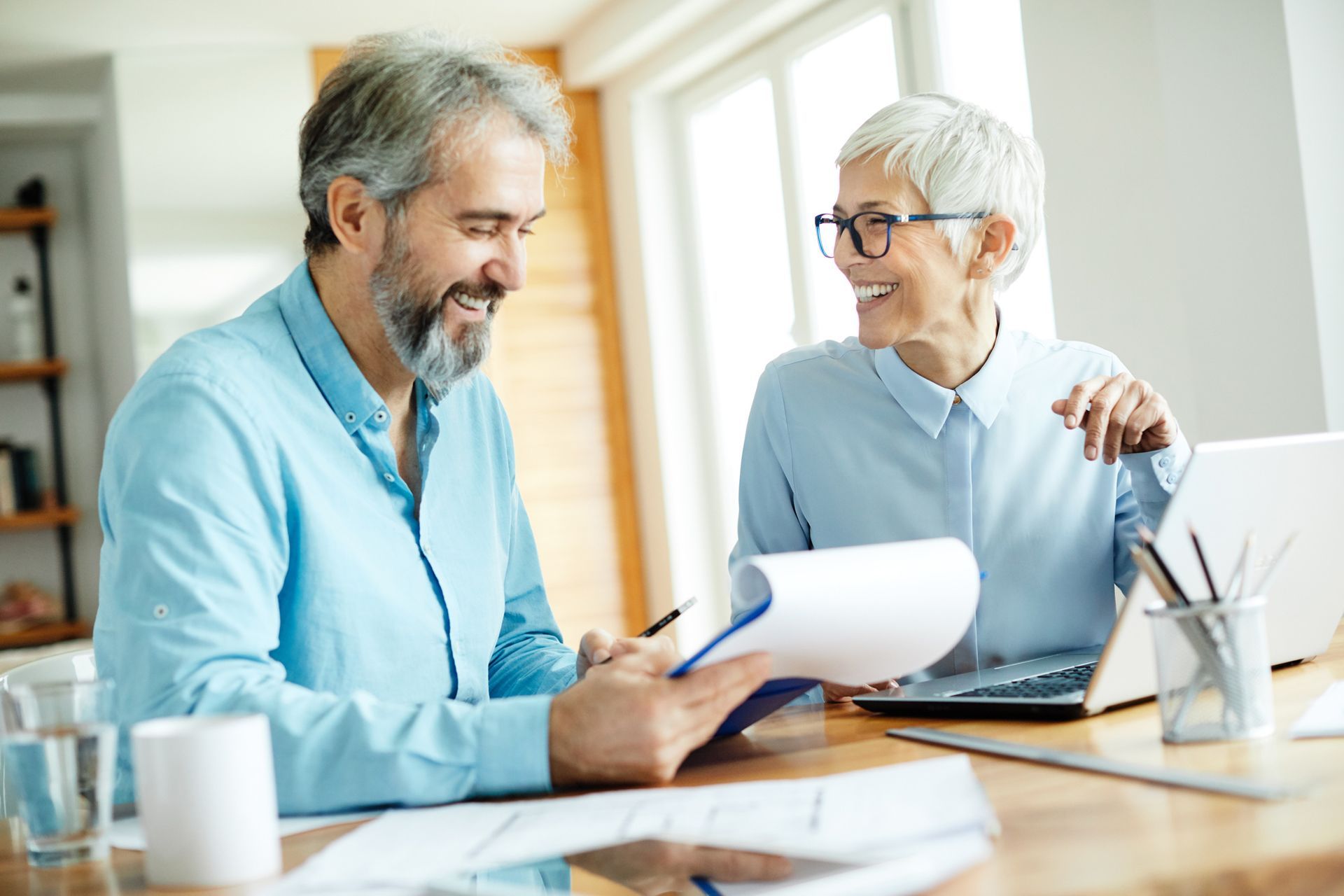 Man and woman smiling, reviewing documents at a table. Laptop and glass of water present.