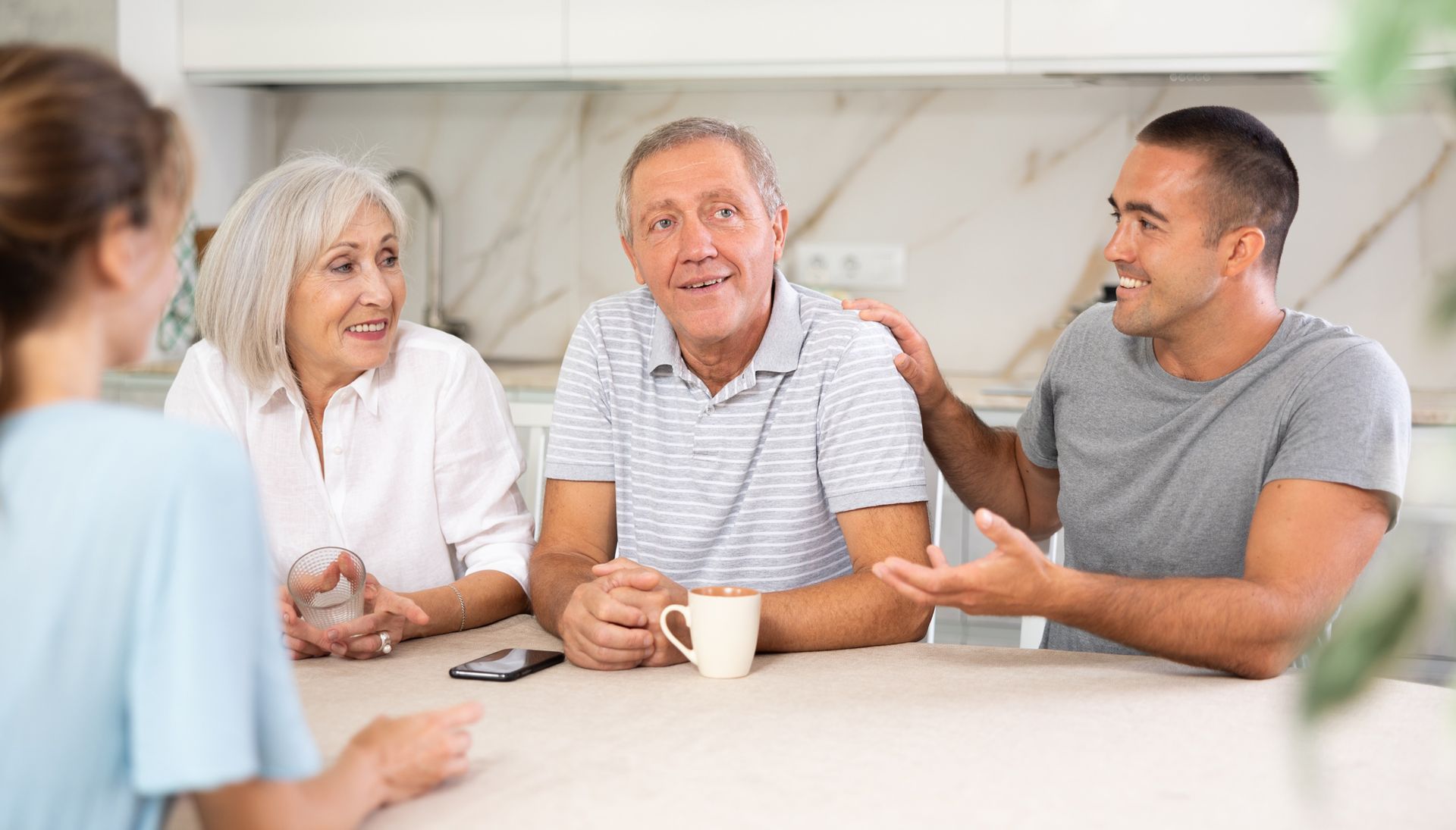 Family at a table: a senior couple, a younger man with hand on the older man's shoulder, and a woman facing them.