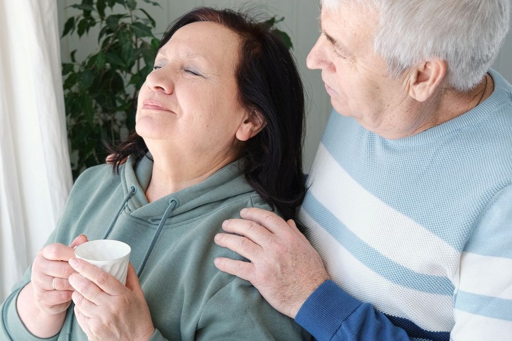 An older woman holds a cup, looking pensive near a window. An older man comforts her with his hand on her shoulder.