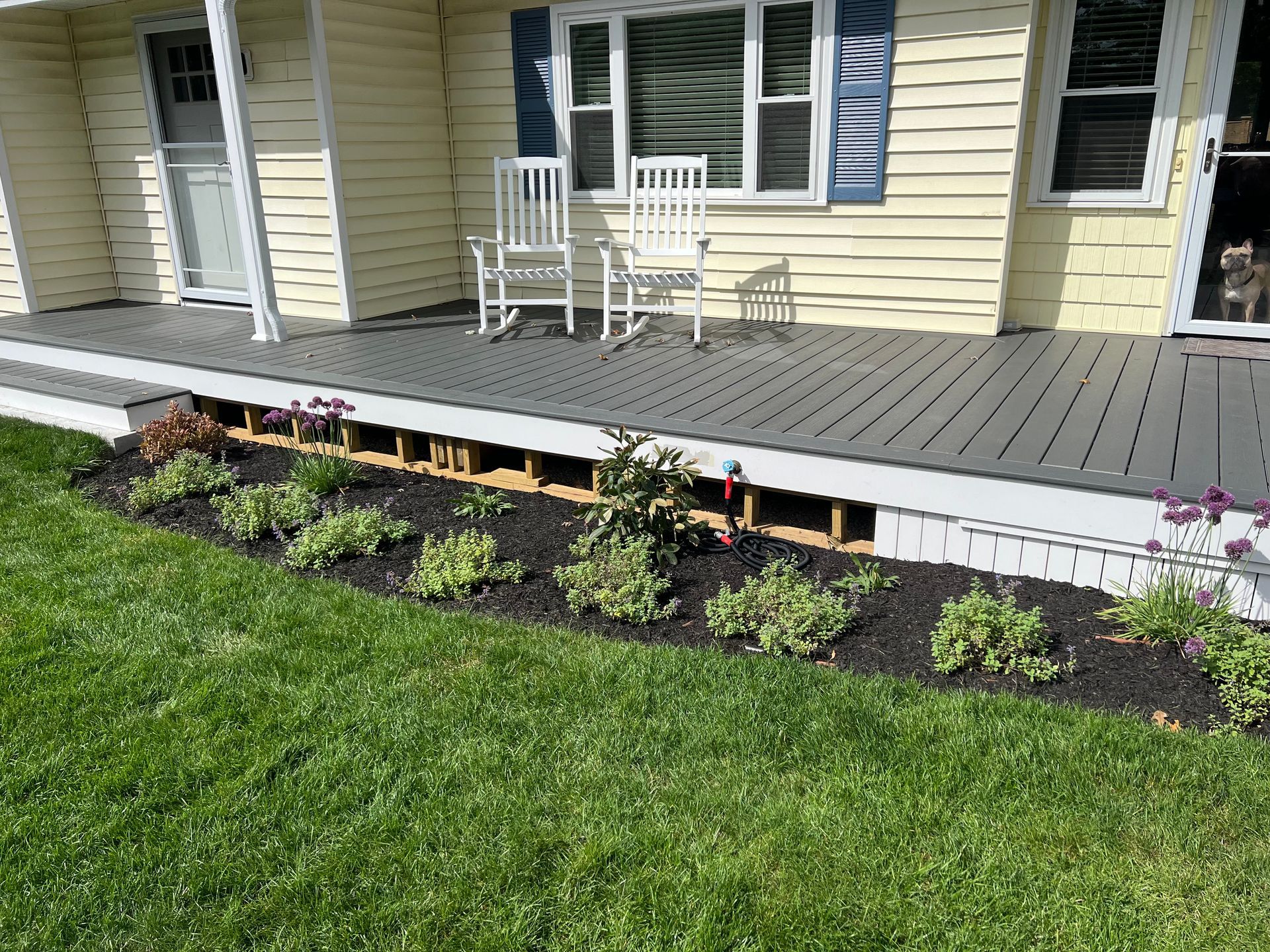 Yellow house with gray porch. Two white rocking chairs sit on the porch. Flower bed with green plants.