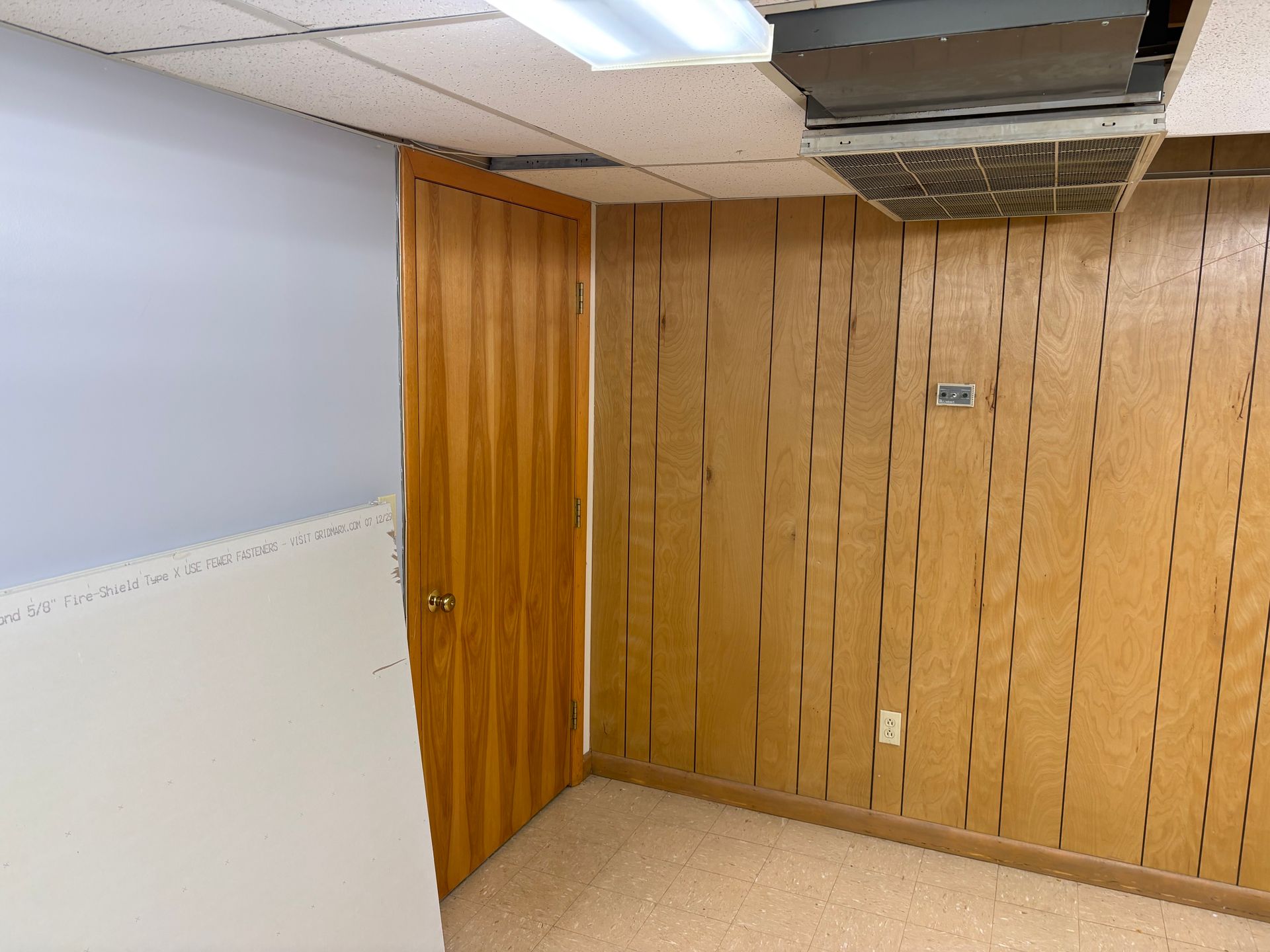 Interior view of a room with wooden paneling, a closed wooden door, and a drop ceiling.