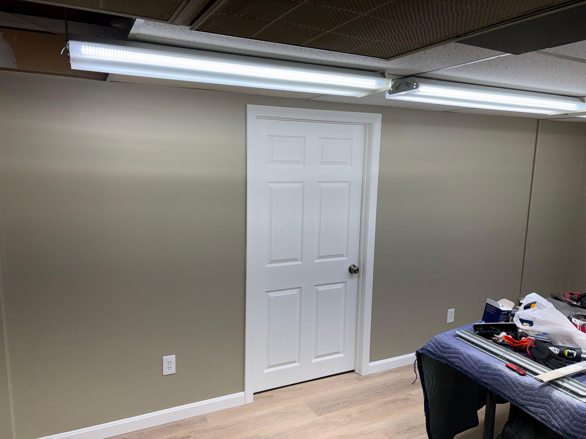 A room with a white door, light-colored walls, and fluorescent lights. A work table is in the foreground.