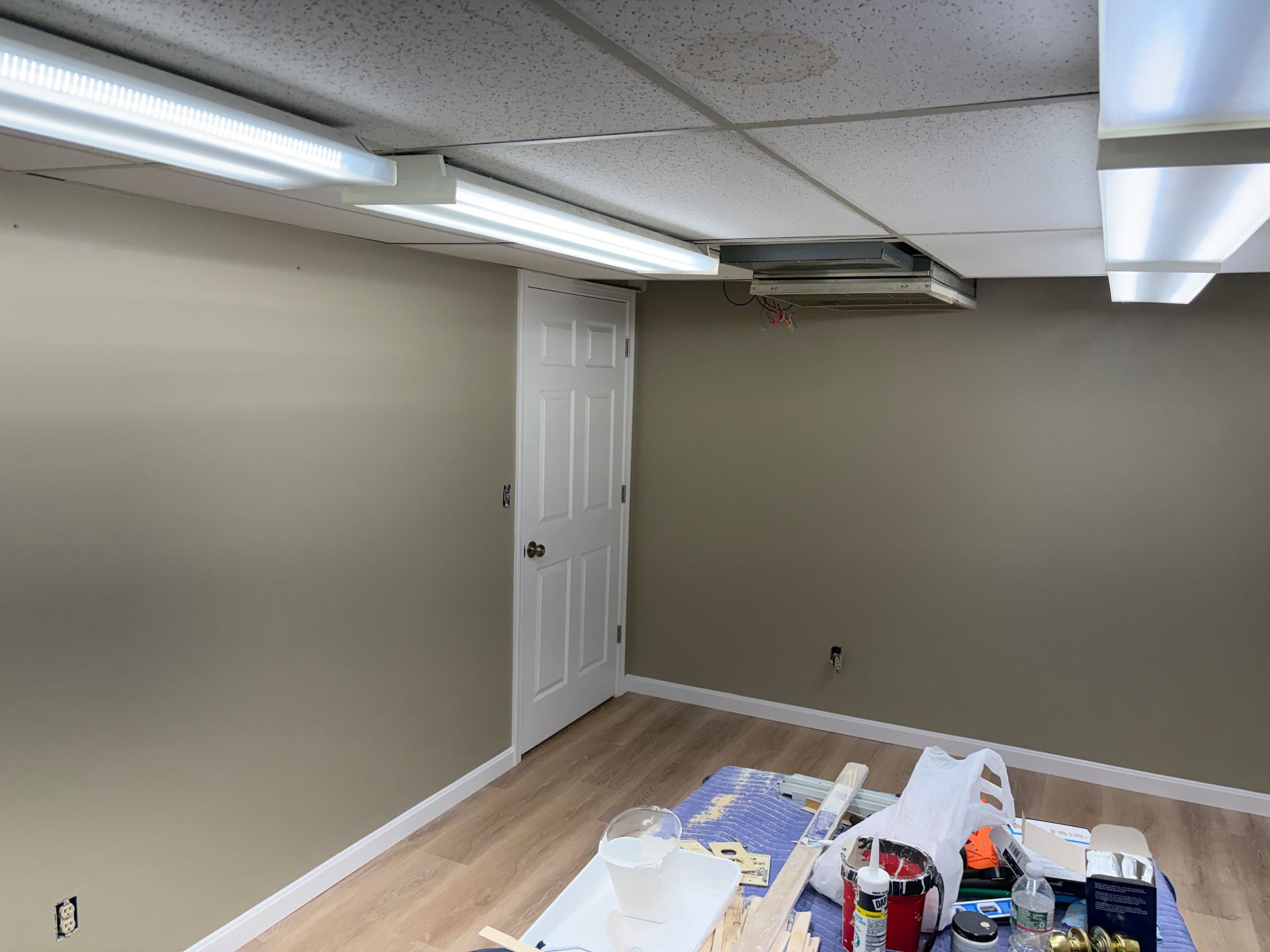 Room with beige walls, fluorescent lights, a white door, and laminate flooring; a work table is in the foreground.