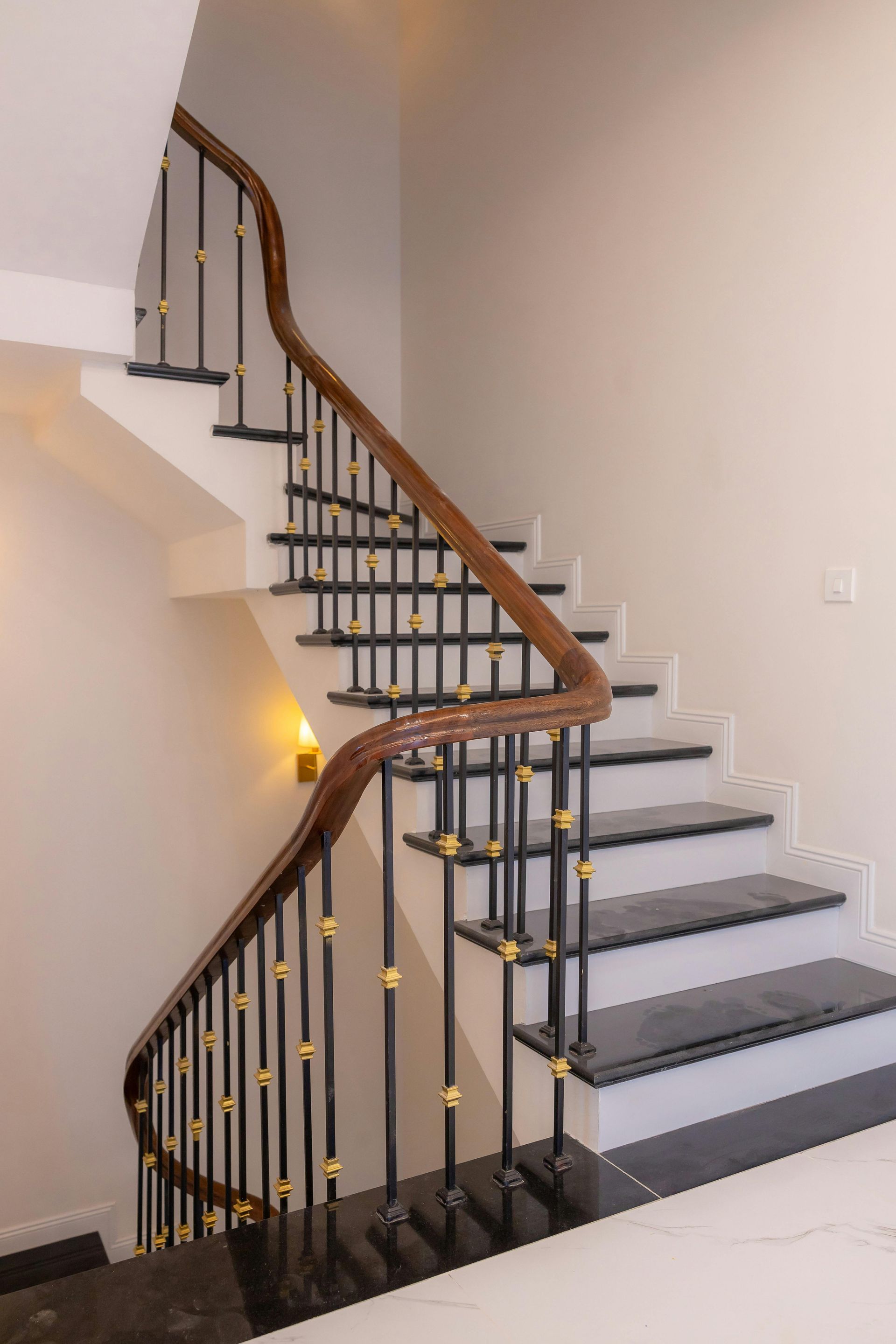 Staircase with wooden handrail, black and gold balusters, and gray steps against a white wall.